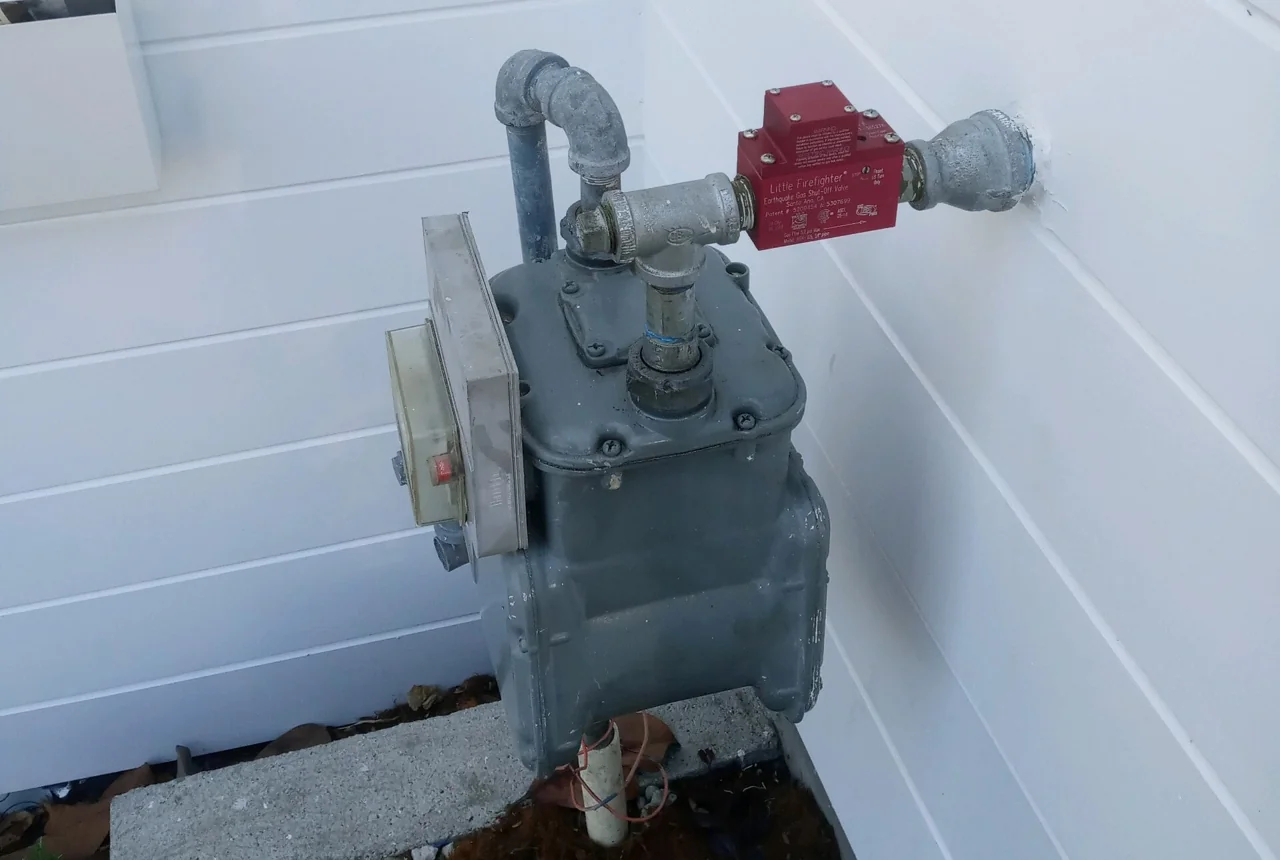 Exterior gas meter and piping with a red shut-off device beside white siding